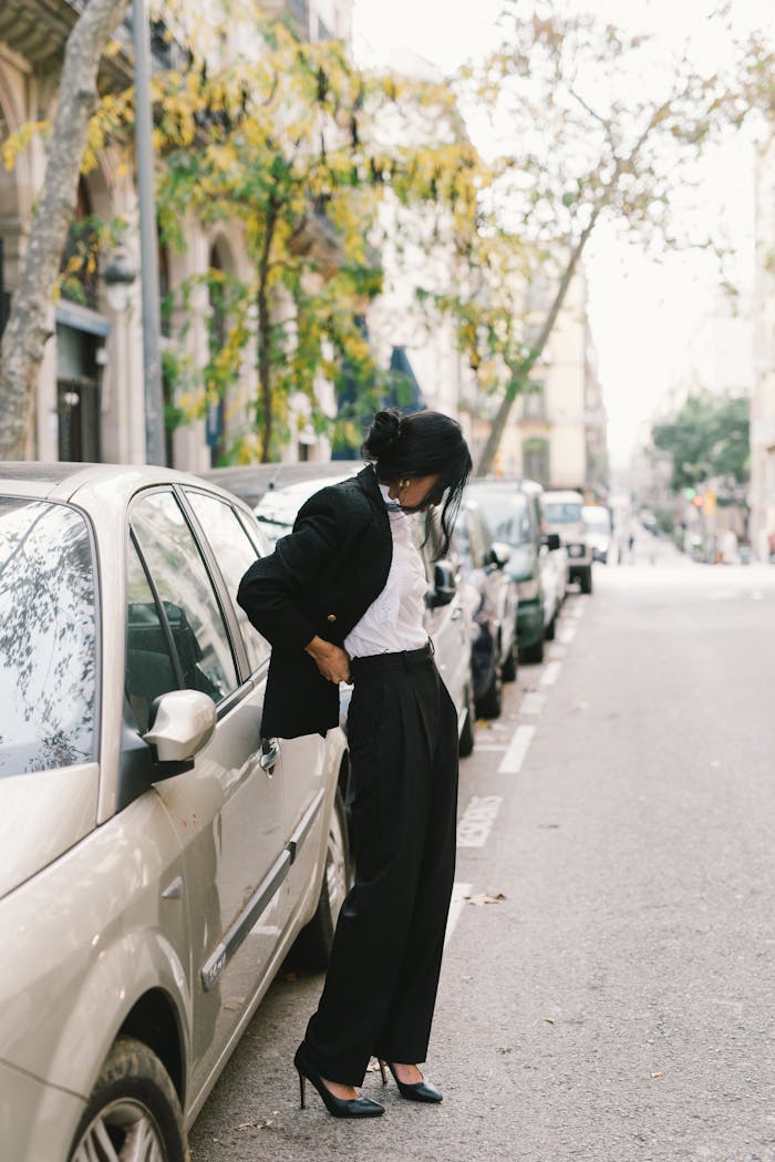 Fashionable woman in a black coat and heels standing by parked cars on a tree-lined street.